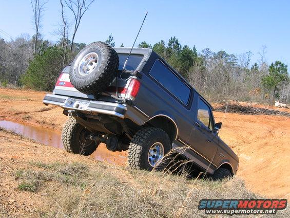 Dark gray Ford Bronco climbing a muddy hill with a spare tire on the back. The vehicle is tilted