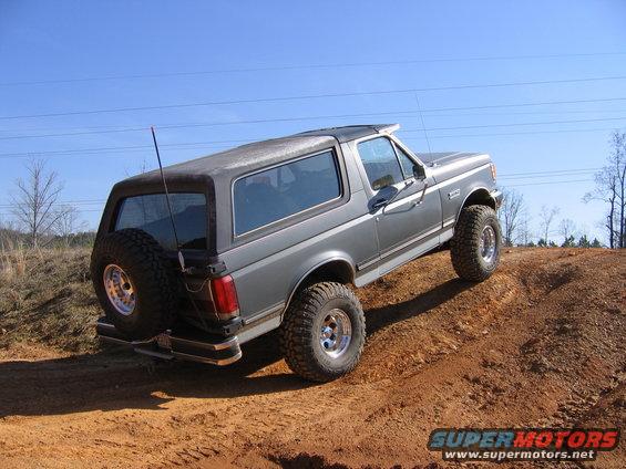 Gray 1989 Ford Bronco with aftermarket wheels and tires driving up a dirt hill. Spare tire mounted on the rear