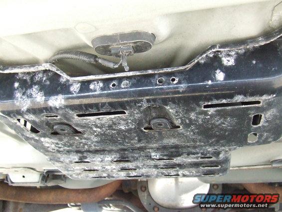 Underside view of a vehicle's underbody, showing a metal shield with holes and some wiring