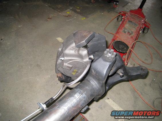 Vehicle axle and brake caliper assembly with brake line, on concrete floor next to red hydraulic jack