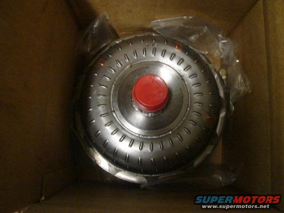Torque converter in a cardboard box, covered in plastic wrap. It has a red cap in the center