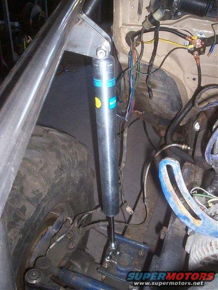 Shock absorber with blue and yellow Bilstein labels, attached to vehicle suspension near a large tire