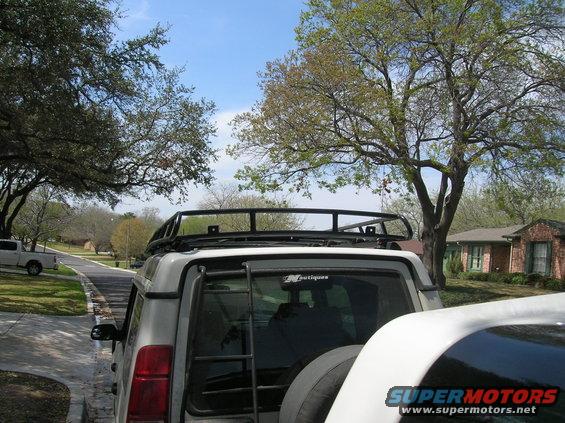 Rear of silver SUV with roof rack and ladder, another white vehicle in foreground