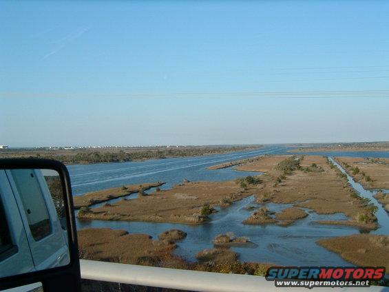 White vehicle side mirror reflecting a marshy waterway under a clear blue sky