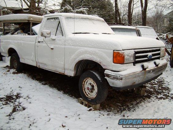 White pickup truck covered in snow, front quarter view. Rusty body, raised on cinder blocks