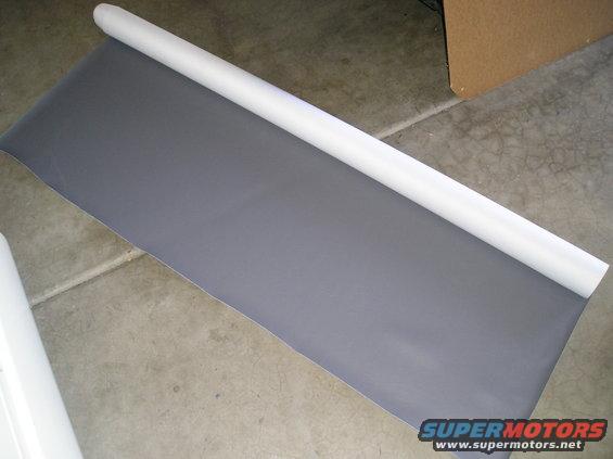 Rolled grey fabric on a white tube, lying on a concrete floor. A cardboard box is visible in the background