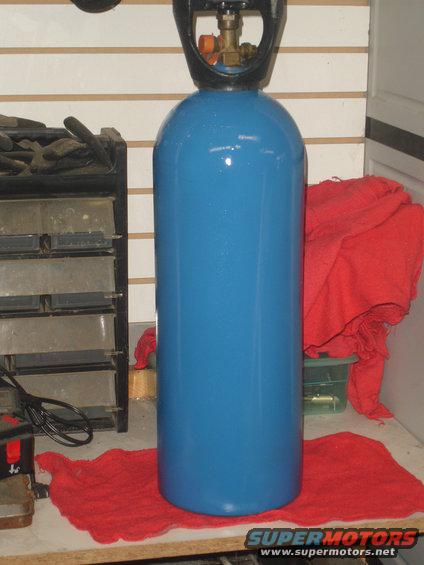 Blue compressed gas cylinder with black regulator on top, resting on a red cloth