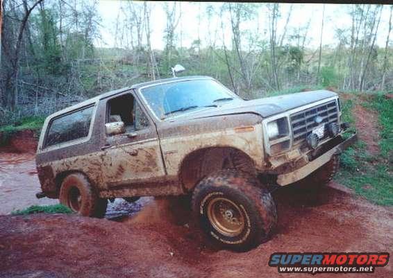 Mud-covered two-tone Ford Bronco driving through dirt and water, front 3/4 view
