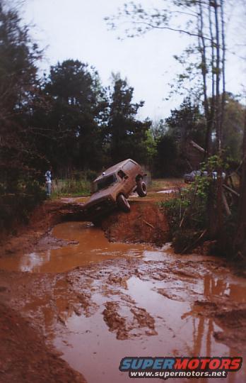 Brown SUV stuck in a deep mud pit on a dirt track, with trees and another vehicle in the background