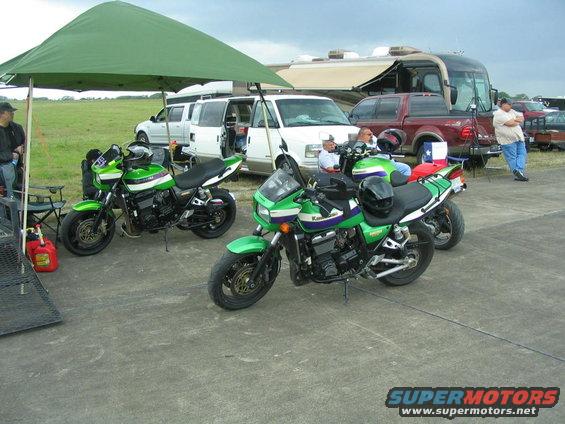 Two green Kawasaki motorcycles parked under a canopy, with vans and an RV in the background