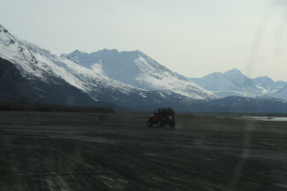 red off-road vehicle driving on a flat, dusty plain with snow-covered mountains in the background