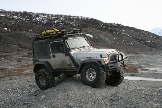 Silver 2003 Jeep TJ Rubicon with roof rack and snorkel parked on gravel with snowy mountains in background