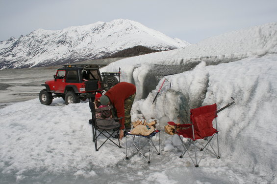 Red Jeep Wrangler parked on snow with a person tending to gear near ice formations and a snowy mountain in the background