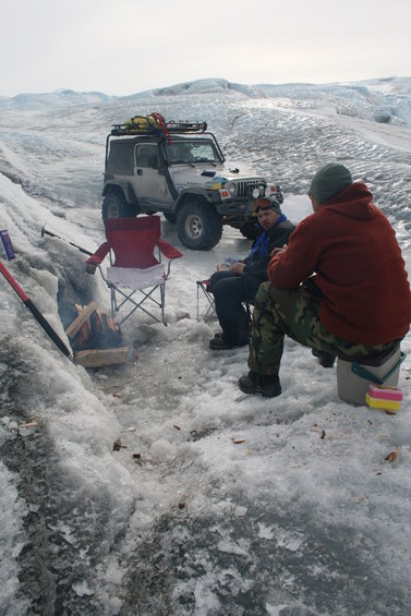 Silver Jeep Wrangler with roof rack parked on ice, two people sitting near a campfire