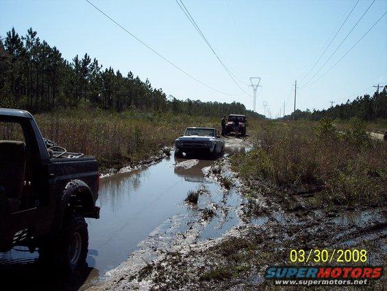 Three off-road vehicles navigating a muddy water-filled trail under a clear sky with power lines overhead