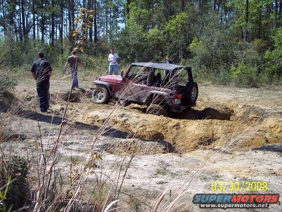 Red Jeep Wrangler partially sunk in a dirt pit with people standing nearby