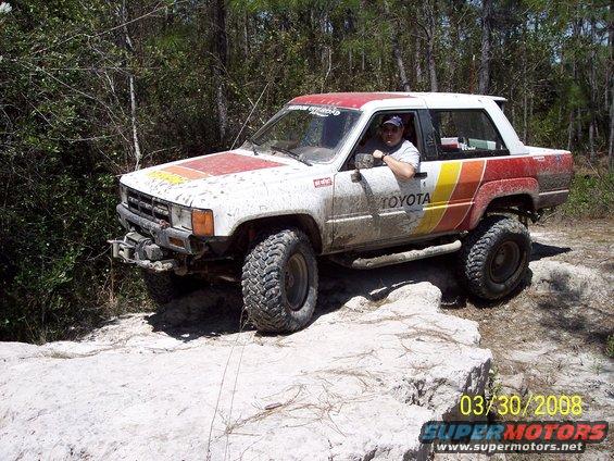 White 1984 Toyota 4Runner with red and yellow stripes on the side, lifted with off-road tires, driving on rocks