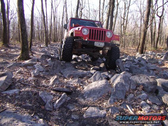 Red 2003 Jeep Wrangler with large tires navigating a rocky trail in a forest