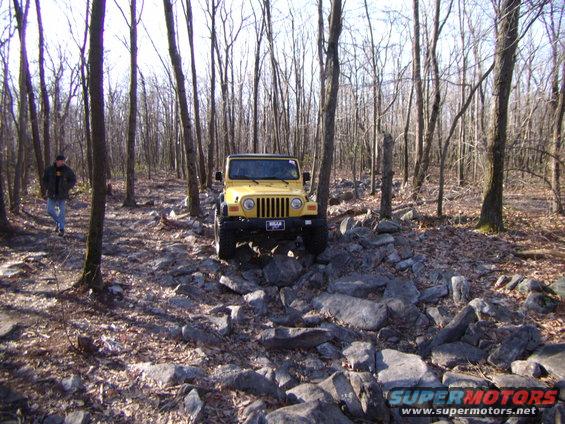 Yellow 2003 Jeep Wrangler on a rocky forest trail, front view. A person walks in the background