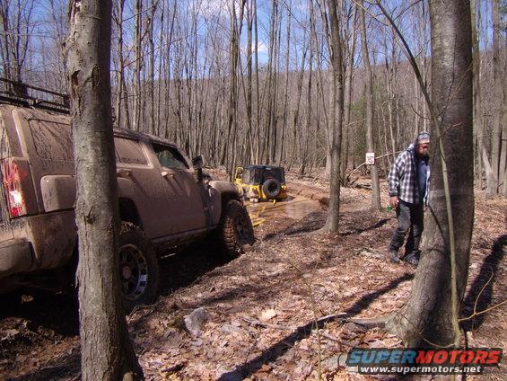 Two off-road vehicles navigating a muddy trail through a forest; one is a tan SUV, the other is a yellow Jeep