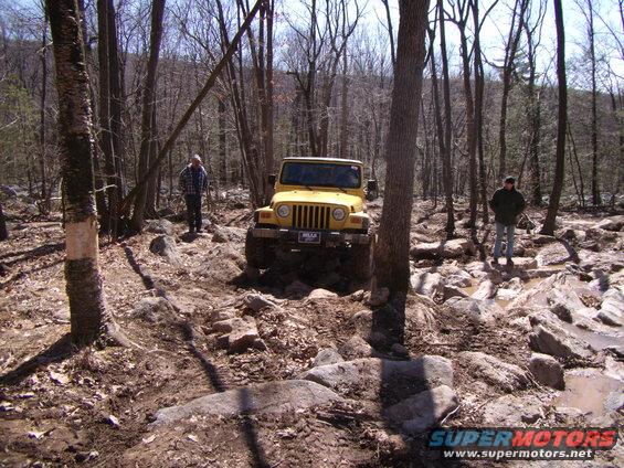 Yellow 2003 Jeep Wrangler navigating a rocky off-road trail with two people standing nearby