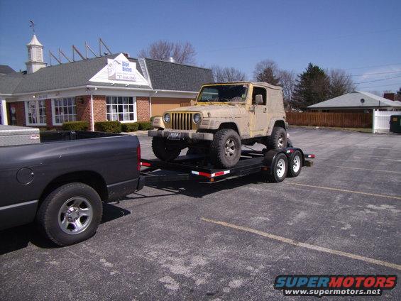 Yellow Jeep Wrangler on a trailer being towed by a dark gray pickup truck in a parking lot