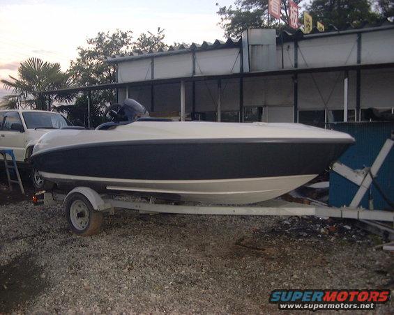 White and dark blue boat on a trailer, parked on gravel near a building. A white SUV is partially visible to the left