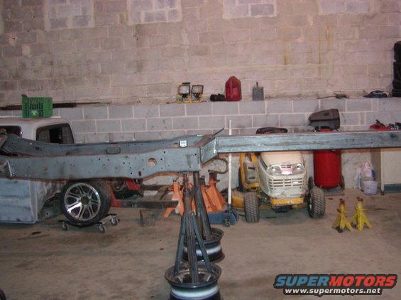 Vehicle frame supported by jack stands in a garage, with a lawn tractor and other equipment nearby