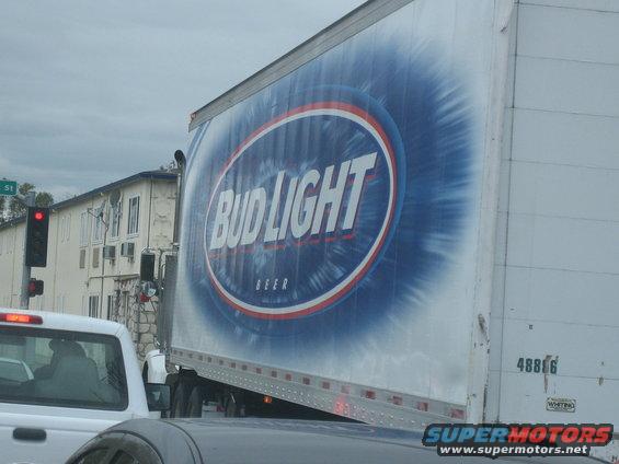 Side view of a Bud Light trailer truck with a white pickup truck in front