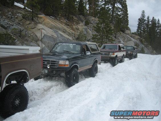 Four Ford Broncos driving on a snow-covered road with rocky hills and trees in the background. The lead Bronco has 'FORD CONSTRUCTION 4X4' on its door