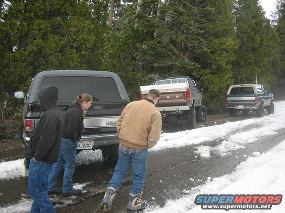 Three pickup trucks and a dark SUV parked on a snowy road with trees in the background