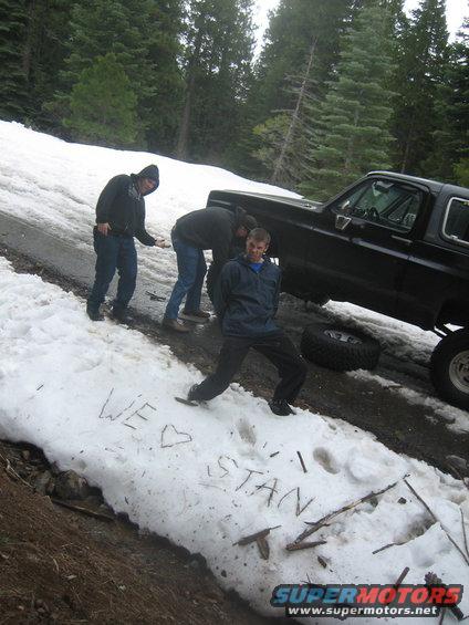 Black SUV with a removed tire in the snow, three people nearby. 'WE <3 STAN' written in snow