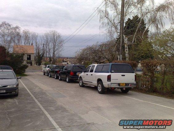 Several trucks and SUVs parked in a row, with a white pickup truck with a camper shell in the foreground
