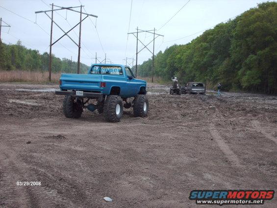 Blue lifted pickup truck with large tires in mud, another truck and ATV in background
