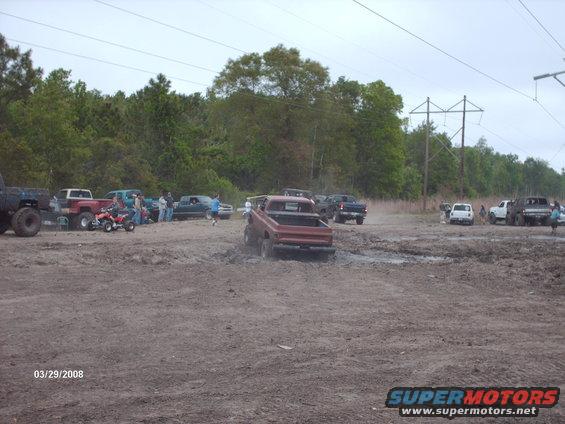 Red pickup truck with lifted suspension driving through mud. The truck has a stepside bed and appears to be from the early 1980s