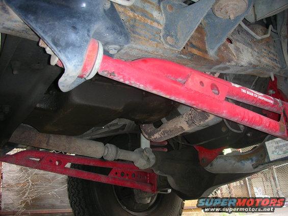 Underside of a vehicle showing red suspension parts and a driveshaft. Text on a red bar reads 'THE REAR OF SUSPENSION'