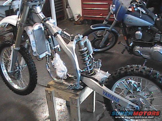Motorcycle frame on stand, front wheel off, rear wheel visible. Another motorcycle in background