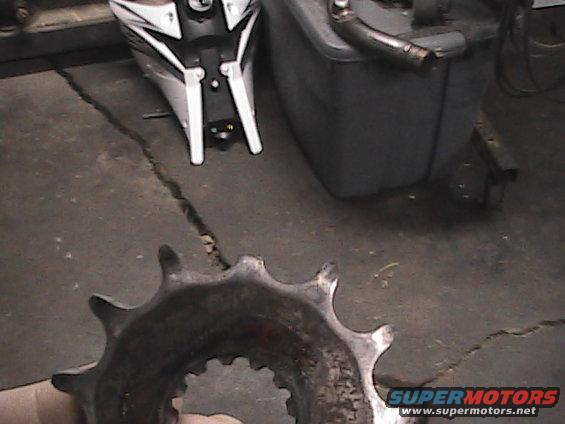 Close-up of a used motorcycle sprocket held in a hand, with a motorcycle body and a grey tank in the background
