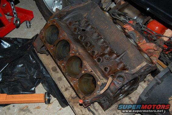 Rusty V8 engine block resting on wooden planks with a black plastic bag and red engine hoist nearby