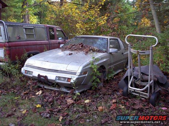 Silver coupe, possibly a Mitsubishi Starion or Chrysler Conquest, overgrown with weeds and leaves on hood. Red pickup truck visible to the left