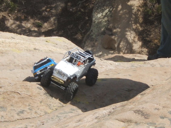 Two remote control rock crawling trucks on a rocky trail. One is white with a roll cage, the other is blue