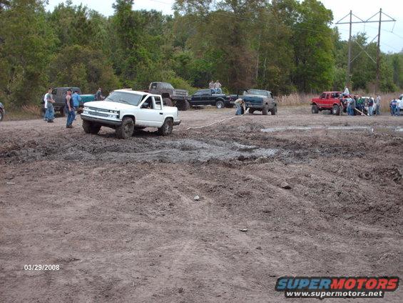 White pickup truck stuck in mud, multiple vehicles and people around