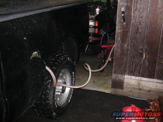 Transferring fuel from a red gas can into a black pickup truck using a hose. Another vehicle is visible in the background