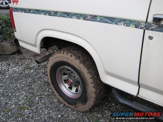 White SUV with camouflage stripe and 4x4 decal on rear quarter panel. Muddy tire and exhaust pipe visible