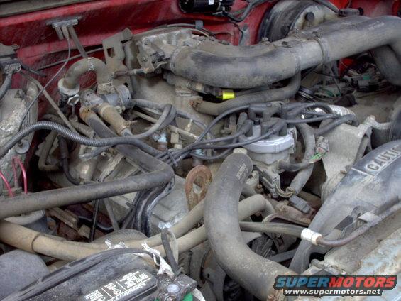Engine bay with hoses, wires, and distributor cap visible. Some text on a battery and a caution label