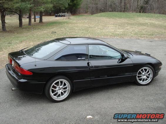 Black 1993 Ford Probe parked on asphalt with trees and a grassy hill in the background