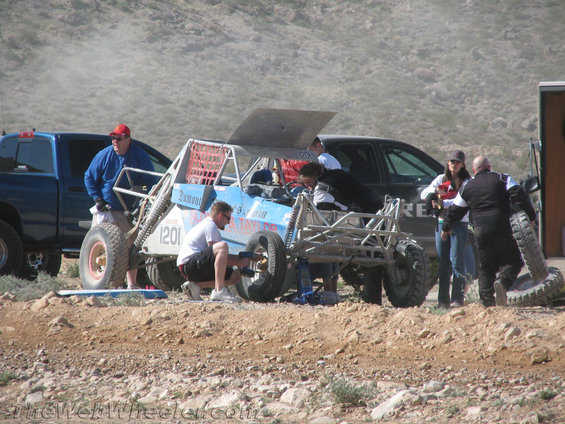 Off-road buggy with number 1201 on side, people working on tire, blue pickup truck nearby