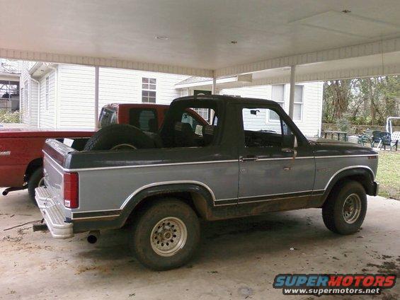 Two-tone gray and black Ford Bronco parked under a carport next to a red pickup truck