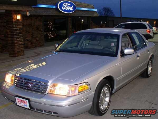 Silver sedan parked in front of a Ford dealership, showing its front grille and headlights