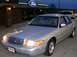 1999 Ford Crown Victoria - A silver sedan is parked outside a dealership with a Ford sign, with another vehicle visible in the background.